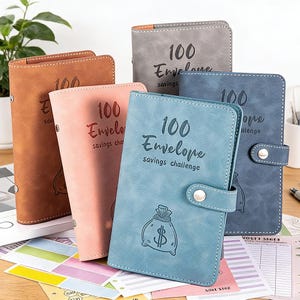 Peut inclure: Collection de cinq classeurs de défis d'épargne en similicuir de différentes couleurs : marron, rose, gris, bleu et marine. Chaque classeur porte l'inscription "100 Envelope savings challenge" et un dessin de sac d'argent.