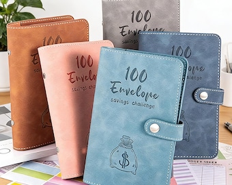 May include: Five faux leather savings challenge binders in shades of brown, pink, grey, blue, and navy. Each binder is embossed with "100 Envelope savings challenge" and a money bag graphic.