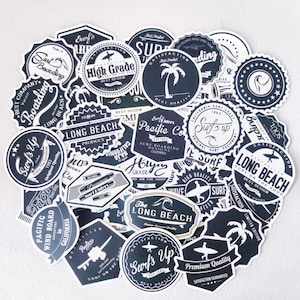 May include: A collection of black and white stickers with various surf-themed designs. The stickers feature text such as "Surf's Up", "Long Beach", "High Grade", and "Pacific Co."