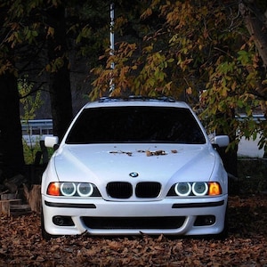Könnte beinhalten: Ein weißer BMW 5er Limousine mit klaren Scheinwerfern und gelben Blinkern. Das Auto steht vor einem Waldgebiet mit heruntergefallenen Blättern auf dem Boden.