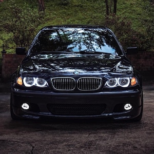 Könnte beinhalten: Ein schwarzer BMW 3er Limousine mit weißen Scheinwerfern und Nebelscheinwerfern. Das Auto steht auf einer gepflasterten Auffahrt.