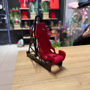 Puede incluir: Un pequeño asiento de carreras rojo con un marco negro, colocado sobre una superficie de madera. El asiento tiene un diseño moldeado con reposacabezas y soportes laterales. El marco negro tiene un diseño geométrico.