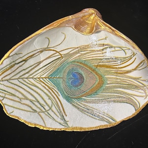 May include: A white seashell with a gold rim and a peacock feather design. The feather is blue, green, and gold.