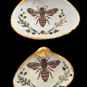 May include: Two white seashells with gold trim, each painted with a detailed image of a bee surrounded by green foliage and small flowers.
