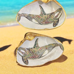 May include: Two seashells with gold trim, each painted with a detailed, colorful orca whale design. The orcas are painted in a paisley pattern with shades of green, red, and gold.