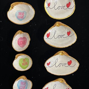 May include: Six small white seashells with gold trim, each with a different Valentine's Day message. The messages include "True Love", "Love", "Be Mine", and "Love" with two red hearts.