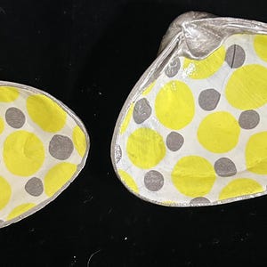 May include: Two seashells painted with a yellow and gray polka dot pattern. The shells are a light gray color and have a smooth, glossy finish.