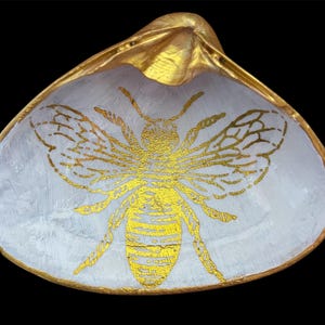 May include: A decorative seashell dish with a gold-painted rim and exterior. The interior is painted white with a gold bee design in the center. The bee's wings and body are detailed with gold paint, creating a striking contrast against the white background.