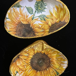 May include: Two golden seashells with a sunflower design. The shells are painted with a white background and feature yellow sunflowers with brown centers.