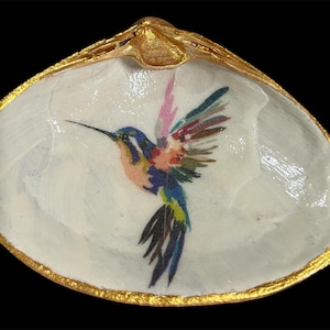 May include: A white seashell dish with a gold-painted rim and a colorful hummingbird illustration. The hummingbird has blue, pink, and yellow feathers. The dish is set against a black background.