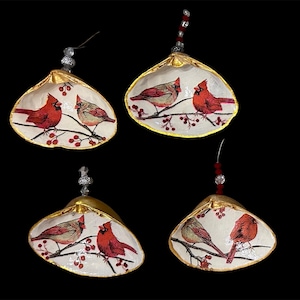 May include: Four seashell ornaments, each featuring a painted scene of two cardinals perched on a branch with red berries. The shells have a gold trim and are suspended by beaded strings, perfect for holiday decor.