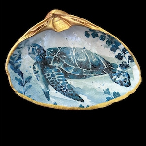 May include: A hand-painted seashell trinket dish featuring a blue sea turtle design. The interior of the shell is painted with a watercolor scene, and the rim is accented with gold paint. The background is black.