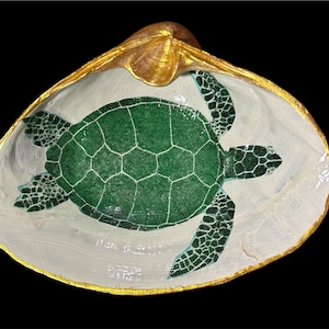 May include: A decorative seashell dish with a painted green sea turtle design. The turtle is centered on a white interior, with a gold-painted rim. The shell is set against a black background.