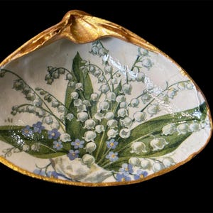 May include: A decorative seashell-shaped dish with a gold-painted rim. The interior features a floral design of white lily of the valley flowers, green leaves, and small blue forget-me-nots against a cream background. The dish is set against a black background.