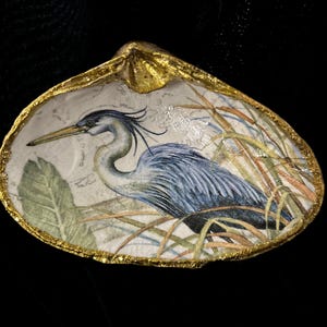May include: A decorative seashell with a gold-painted rim. The interior features a detailed illustration of a blue heron in a marsh setting, with reeds and foliage. The heron is depicted with blue and gray feathers.