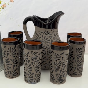 May include: A ceramic pitcher and six matching glasses. The set is gray with black floral designs and black rims. The interior of the glasses and the pitcher's interior are brown. The pitcher has a black handle.