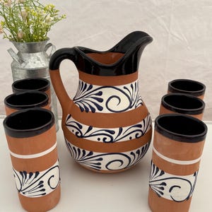 May include: A terracotta pitcher and six matching tumblers featuring a smooth, unglazed clay finish with glossy black rims and handles. The set is decorated with horizontal white bands containing intricate, hand-painted dark blue floral scrollwork.