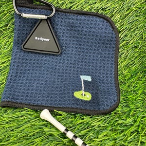 May include: Navy blue golf towel with a black trim, a black carabiner clip, and a triangular tag that reads "Batiyeer." The towel has an embroidered golf flag design. Two white golf tees with black stripes are in the foreground.
