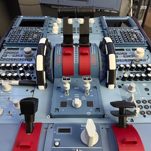 May include: A flight simulator throttle quadrant featuring dual black throttle levers with red accents, engine start switches labeled ENG 1 and ENG 2, and a parking brake lever labeled PARK BRK. The control panel includes various knobs and switches.