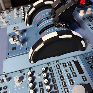 May include: A light blue flight simulator throttle quadrant featuring two large black and white trim wheels, a black thrust lever with a red button, and various control panels with labeled buttons and knobs including ENG 1, ENG 2, NAV, VOR, LS, and GLS.