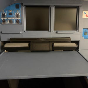 May include: A grey aircraft cockpit flight deck panel featuring two blank rectangular display screens, a retractable tray table, and various control knobs labelled with text including LOUD SPEAKER, CONSOLE/FLOOR, and FOOT WARMER, plus an American Airlines logo.
