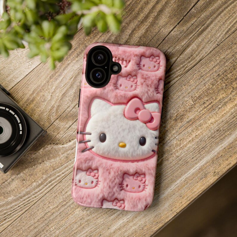 May include: Pink phone case featuring a textured design with a large Hello Kitty face and multiple smaller Hello Kitty images. The case has a raised bow and a camera cutout.