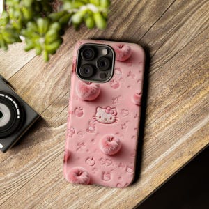 May include: Pink phone case featuring a Hello Kitty design with embossed details and fuzzy apple accents. The case has a camera cutout and is decorated with various embossed shapes. The case is on a wooden surface.