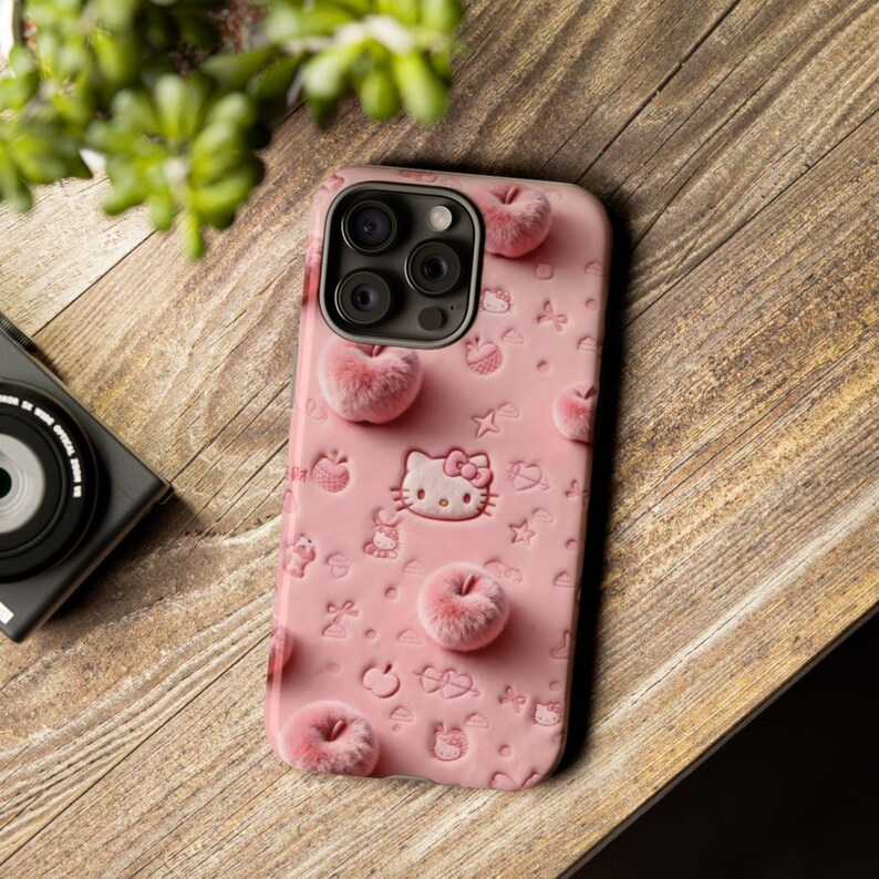 May include: Pink phone case featuring a Hello Kitty design with embossed details and fuzzy apple accents. The case has a camera cutout and is decorated with various embossed shapes. The case is on a wooden surface.