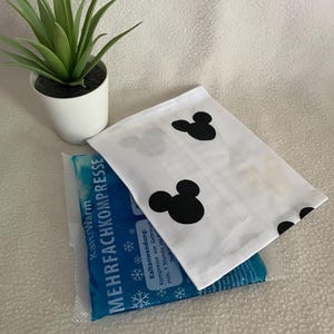 Könnte beinhalten: Eine weiße Stofftasche mit schwarzen Mickey Mouse Ohren bedruckt. Die Tasche ist über eine blaue und weiße Kühlkompresse mit dem Text "Kalt/Warm MEHRFACHKOMPRESSE" und Gebrauchsanweisung gefaltet.