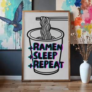 以下が含まれることがあります： ラーメンのカップと箸、麺の白黒イラストが描かれたフレーム入りプリント。 「RAMEN SLEEP REPEAT」の文字は、シアンとマゼンタのアクセントで3D効果で印刷されています。 フレームは明るい茶色です。