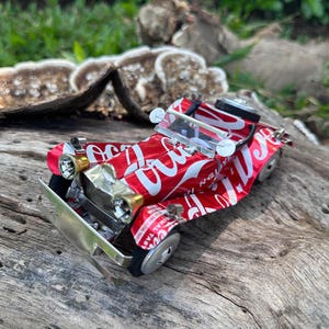 May include: A miniature car crafted from recycled Coca-Cola cans, showcasing a red and white design. The model car has detailed features such as a silver bumper, headlights, and wheels. A creative example of upcycling, this is a unique piece of art.