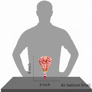 Puede incluir: Un peque&ntilde;o globo aerost&aacute;tico rojo y blanco con el logotipo de Coca-Cola, que mide 15 cm de alto y 10 cm de ancho. El globo tiene detalles dorados y una base roja.