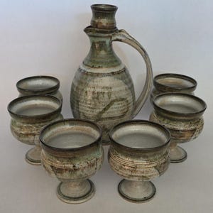 May include: A ceramic beverage set consisting of one pitcher with a handle and six matching stemmed goblets. Each piece features a mottled glaze in earthy tones of beige, brown, and muted green with a textured, speckled finish throughout.