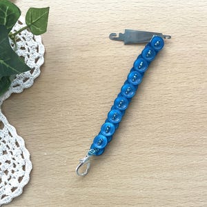May include: A blue button bracelet with a silver clasp and a metal tool. The bracelet is made of small, round, blue buttons strung together. The metal tool is silver and has a hook shape.