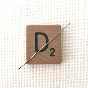 May include: A wooden Scrabble tile with the letter "D" and the number "2" printed in black. A silver needle is diagonally across the tile. The tile is brown and sits on a white, textured surface.