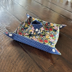 May include: A fabric catchall tray with a floral pattern interior and a navy blue polka dot exterior. The tray is held together with white snaps. The tray is open and ready to hold small items.