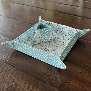 May include: A fabric tray and a smaller, triangular container. The tray is light blue with white polka dots on the outside and a floral pattern on the inside. The container has the same floral pattern. The tray is folded up at the corners.