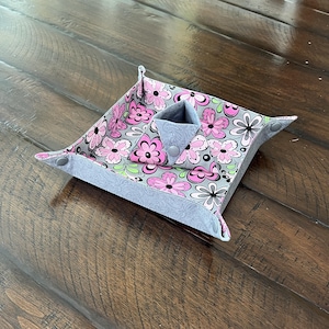 May include: A fabric catchall tray with a floral pattern in pink, gray, and black. The tray has a gray border and a small, triangular pocket with snap closures. The tray is designed to hold small items.