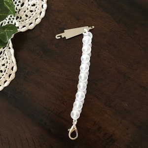 May include: A silver-toned button bracelet with a gold-colored clasp. The bracelet is made of white, round buttons strung together. A silver-toned metal piece is attached to the top of the bracelet.