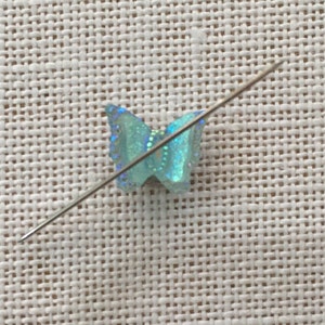 May include: A small, light blue butterfly-shaped embellishment with iridescent glitter accents, pierced by a silver needle. The butterfly is positioned on a textured, off-white fabric, suggesting a craft or sewing project.