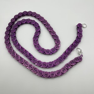 May include: A purple button strap with silver-colored clasps. The strap is made of small, round, purple buttons strung together. The buttons are a solid, shiny purple color. The clasps are attached to the ends of the strap.