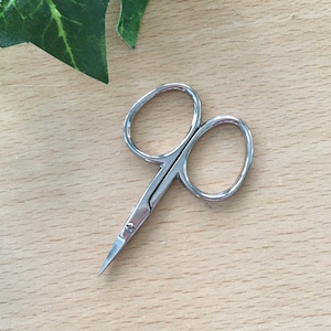 May include: Small, silver-colored scissors with rounded finger holes and sharp, pointed blades. The scissors are designed for precision cutting and are likely used for grooming or crafting. The image is taken from a close-up angle.