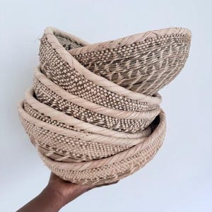 May include: A stack of four woven baskets made of natural materials. The baskets are beige and have a woven pattern. The baskets are stacked on top of each other and are being held in someone's hand.