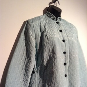 May include: A light blue quilted jacket with black buttons and trim. The jacket has a high collar and long sleeves. The fabric has a subtle pattern. The cuffs and collar are trimmed with a black, decorative design. The jacket is displayed on a black mannequin.