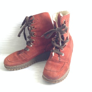 May include: A pair of rust-colored suede lace-up boots featuring dark brown laces, gold-toned metal eyelets, and a thick, textured brown rubber sole. The interior of the boots is lined with a light-colored, fuzzy material visible at the top opening.