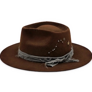 Pode incluir: Chapéu fedora castanho com aba larga e faixa de tecido cinza. O chapéu é decorado com um desenho de constelação de pequenas contas prateadas e uma estrela dourada. O chapéu é feito de material semelhante a feltro. A aba tem cerca de 10 cm de largura.
