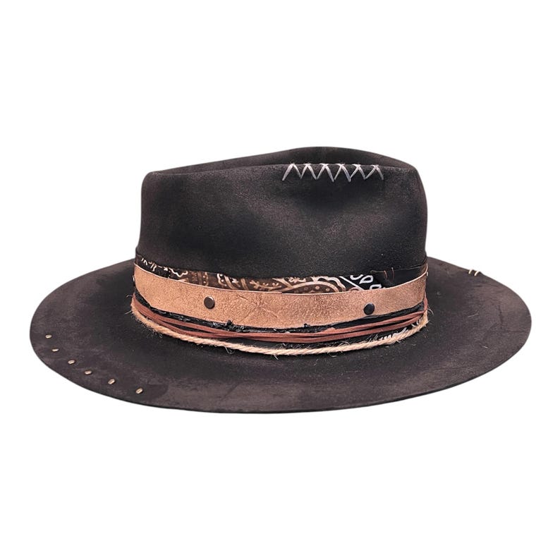 May include: A black fedora hat with a wide brim. The hat has a decorative band with a paisley pattern, leather accents, and a rope trim. Silver studs are placed around the brim. White stitching accents the top of the hat.