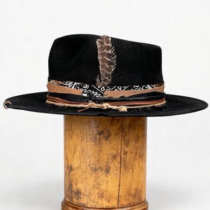 May include: A black fedora hat with a wide brim, featuring a patterned band, leather details, and a decorative feather. The hat is presented on a wooden stand, highlighting its fashionable design.
