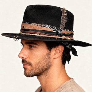 May include: Black fedora hat with a wide brim, decorated with a patterned band, leather details, and a feather. The hat has a rope band and two gold-coloured rings on the brim. A black and white bandana is tied around the hat.