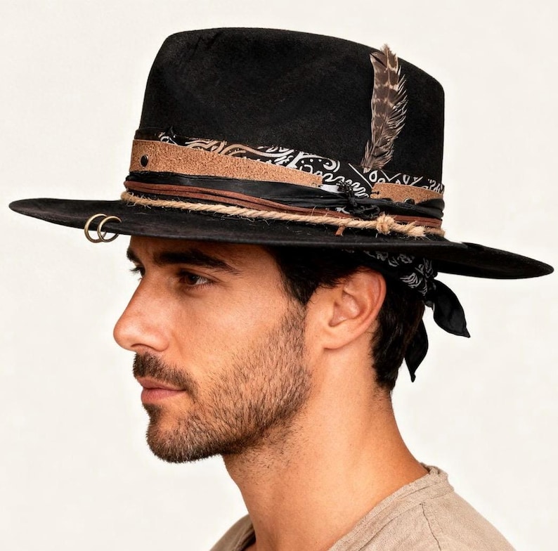 May include: Black fedora hat with a wide brim, decorated with a patterned band, leather details, and a feather. The hat has a rope band and two gold-coloured rings on the brim. A black and white bandana is tied around the hat.
